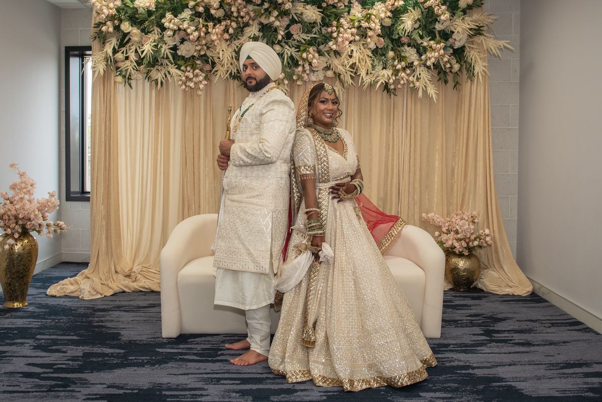 A South Asian bride and groom in traditional attire stand back-to-back near a floral arch in an elegant indoor setting.