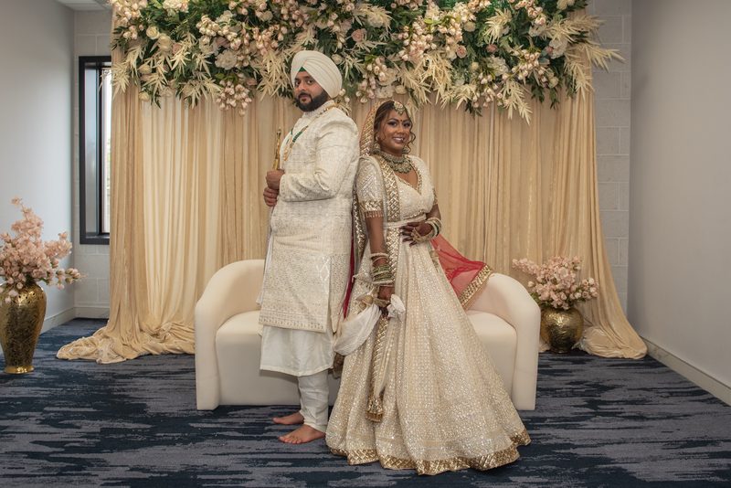A South Asian bride and groom in traditional attire stand back-to-back near a floral arch in an elegant indoor setting.