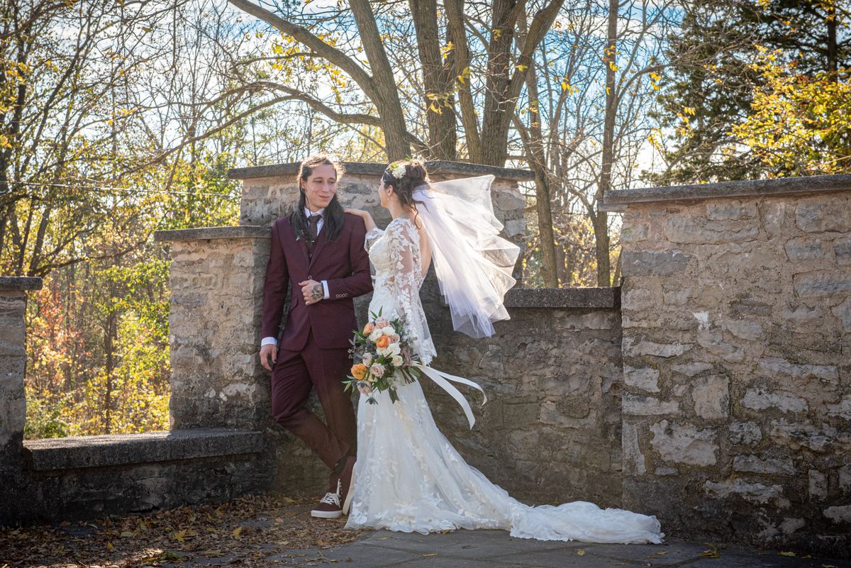 A bride and groom stand by a stone wall in an autumn garden, illuminated by golden sunlight through bare trees.