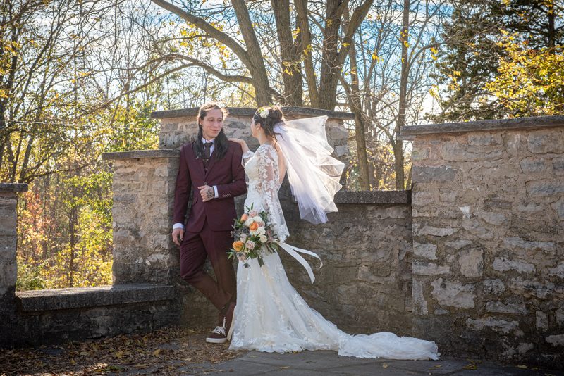 A bride and groom stand by a stone wall in an autumn garden, illuminated by golden sunlight through bare trees.