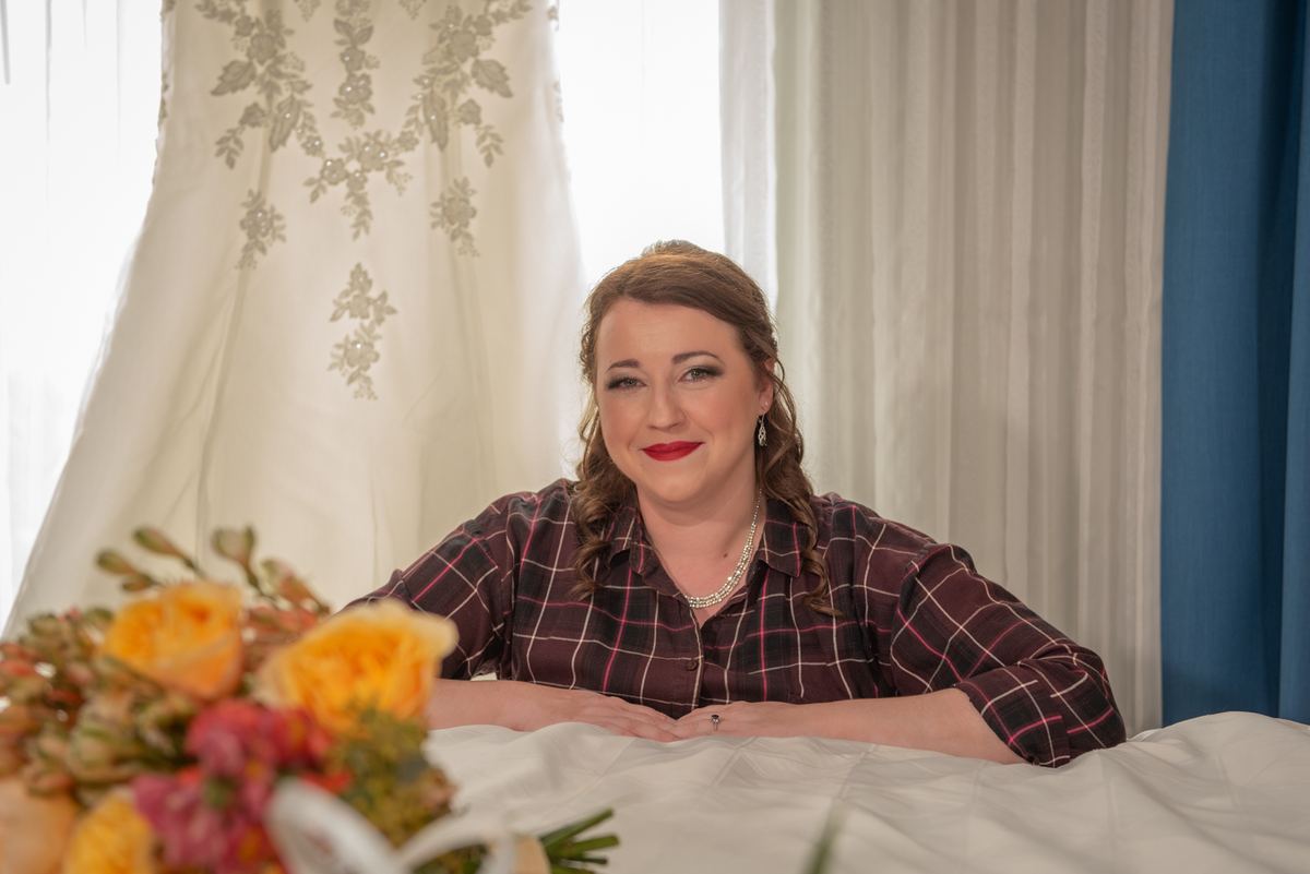 A smiling bride sits at a table with flowers, her wedding gown hanging behind her on sheer curtains.