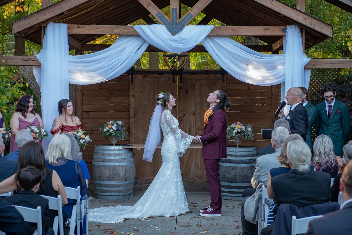 A bride and groom exchange vows under a wooden arbor draped with white fabric during an outdoor ceremony.