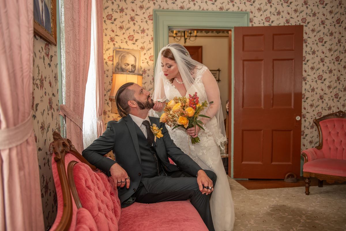 A bride in a white gown and bouquet leans over a smiling groom in a dark suit on a pink velvet sofa.