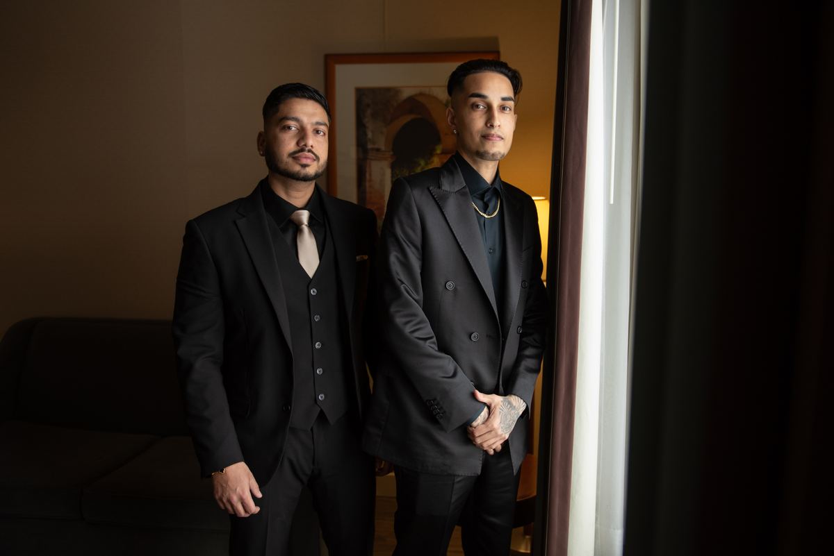 Two men in black suits stand thoughtfully by a bright window, their faces and suits highlighted by dramatic sidelight.