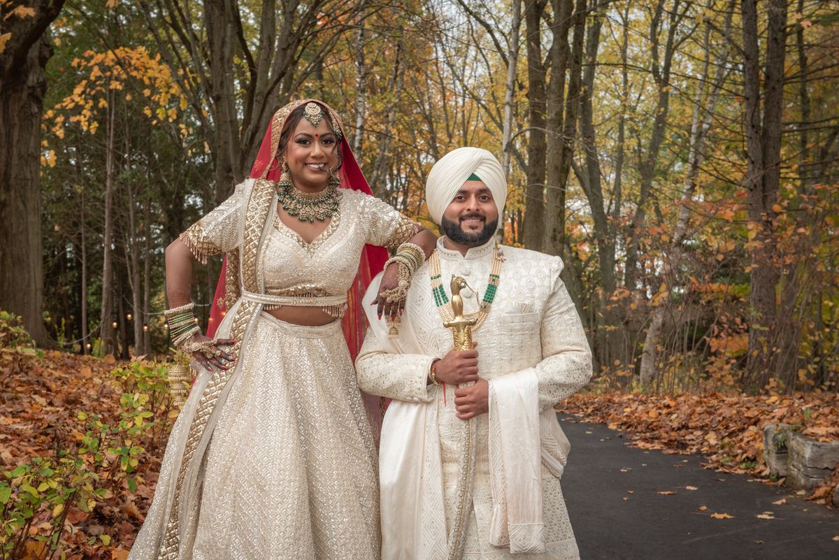 A smiling South Asian bride and groom pose in an autumn forest, surrounded by golden leaves and soft daylight.