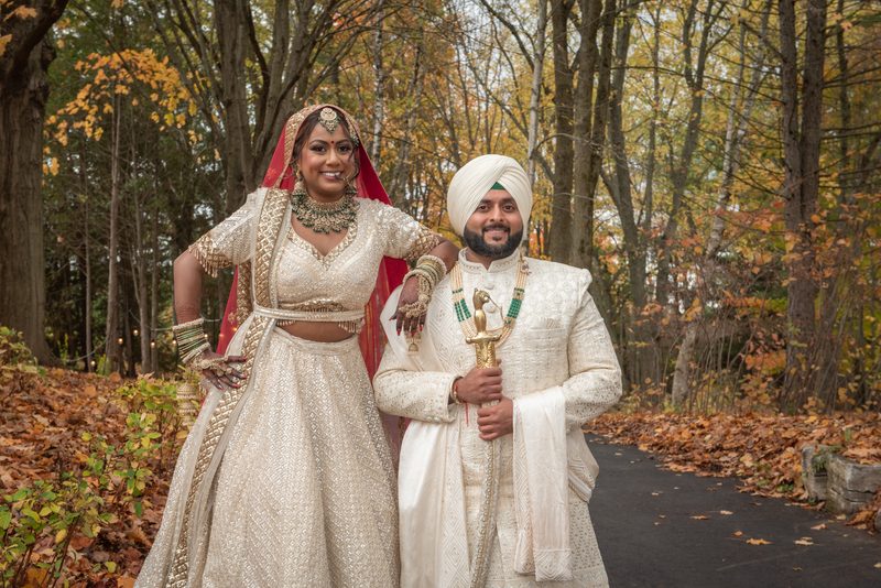 A smiling South Asian bride and groom pose in an autumn forest, surrounded by golden leaves and soft daylight.