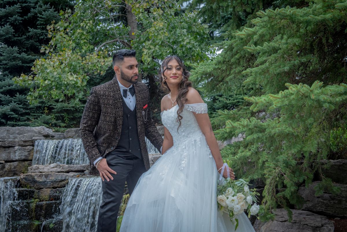 A bride in a white gown and groom in a brown jacket pose beside a small stone waterfall surrounded by lush greenery.