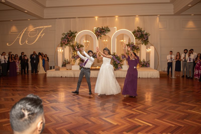 Fazana and Aman joyfully dancing on the polished wooden floor at their wedding reception, surrounded by applauding guests.
