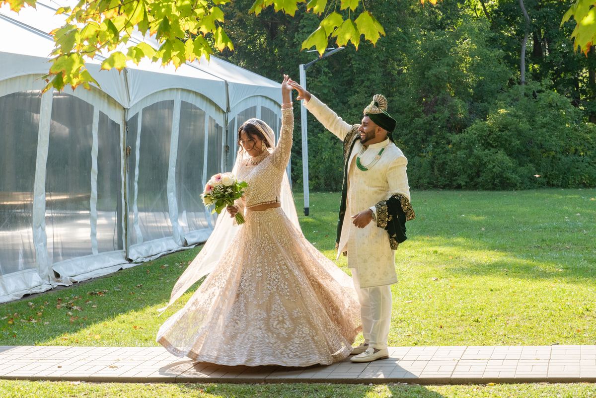 Bride in an embellished lehenga and groom in sherwani dance outdoors, hand in hand, under golden autumn leaves.