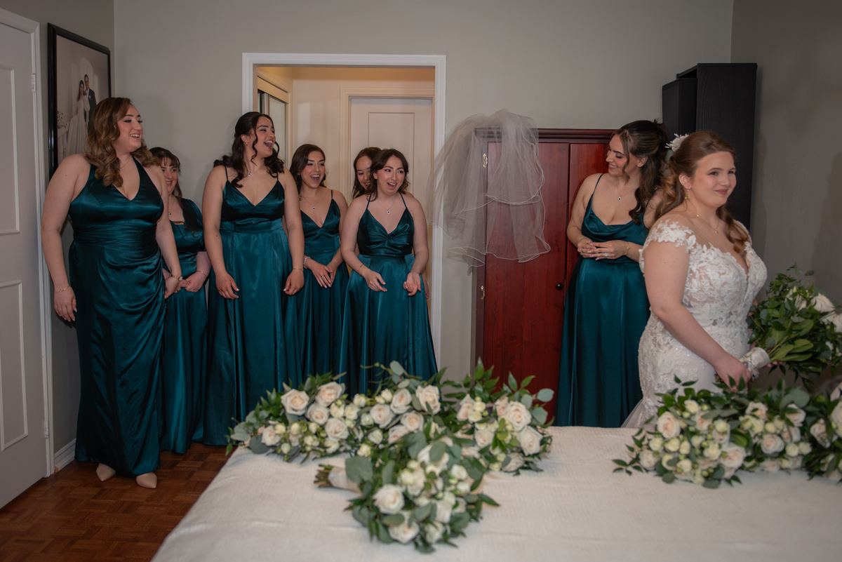 A bride in a white lace gown stands by a table with white roses as her bridesmaids look on, smiling.