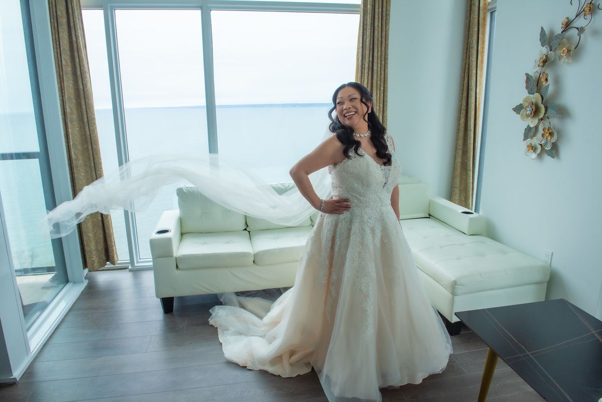 A smiling bride in a modern room with floor-to-ceiling ocean views, her veil fluttering in the breeze.