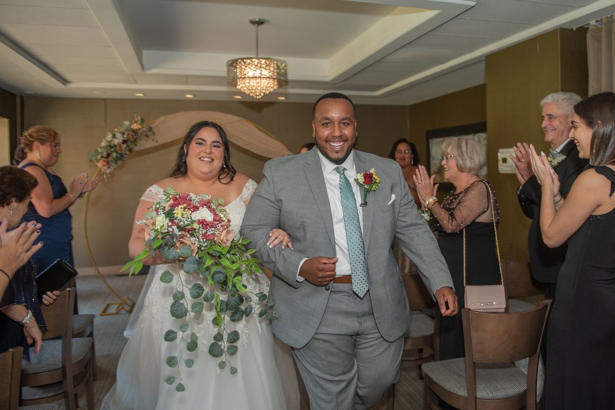Bride and groom walk down the aisle hand-in-hand, smiling as guests clap, under a floral arch and chandelier lighting.