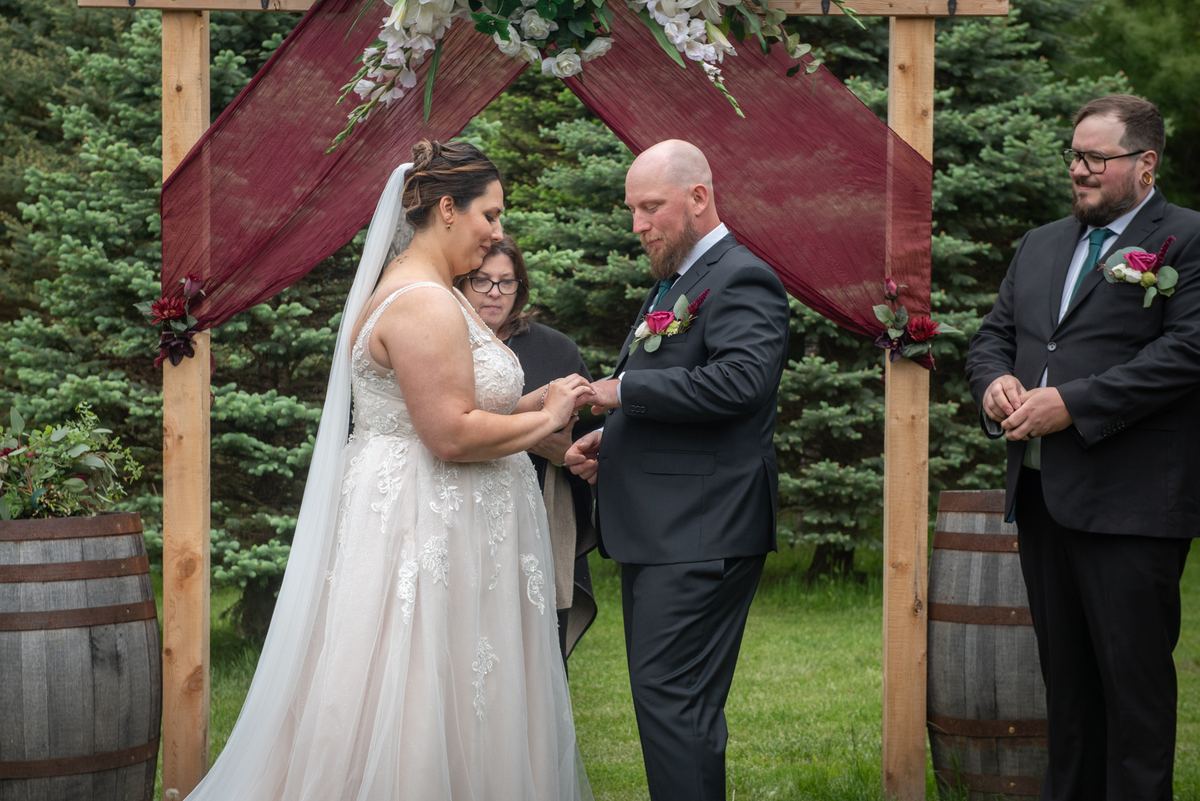 Bride and groom exchange rings under wooden arch with burgundy fabric and flowers, surrounded by trees.