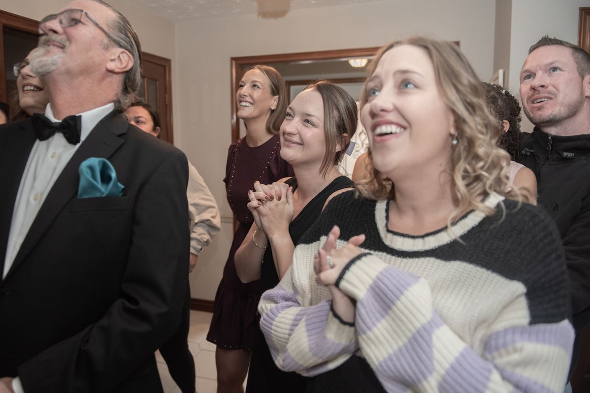 Guests in formal attire smile and gaze upward with joy during a wedding celebration, their faces illuminated by warm lights.