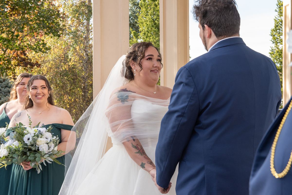 Bride in white gown and veil smiles at groom in navy suit as they exchange vows under filtered sunlight.