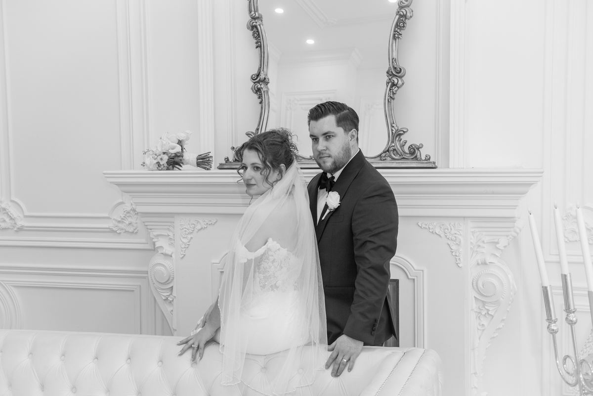 Bride in lace gown and veil with groom in tuxedo beside ornate sofa, illuminated by soft light in classic room.