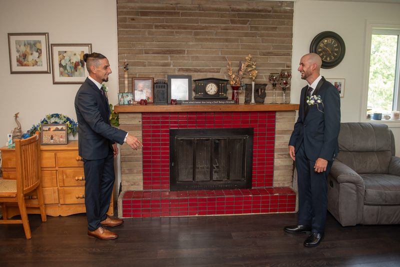 Two grooms in formal suits smile and gesture towards each other in a living room with a red-tiled fireplace.