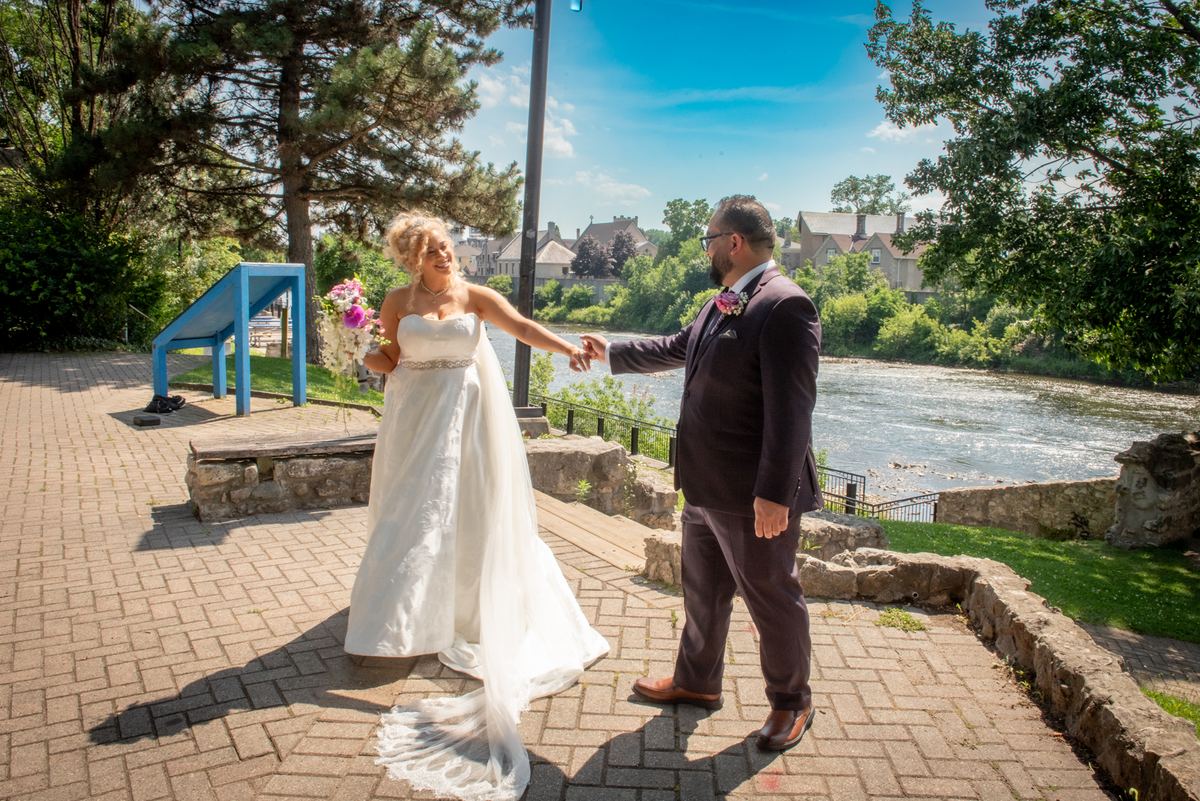Bride and groom stand hand-in-hand on a stone-paved riverside walkway, smiling joyfully under sunny skies.