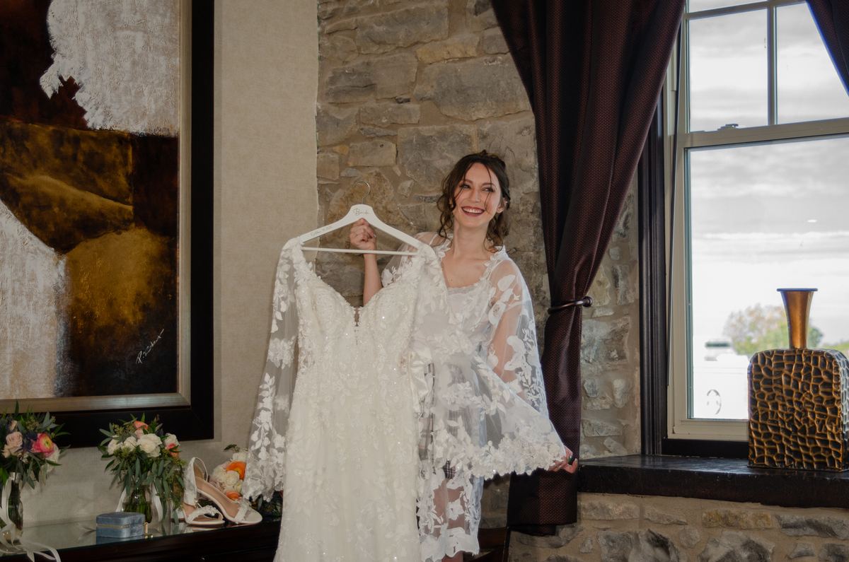 A smiling bride in a lace robe holds up her white wedding gown, illuminated by natural light in a stone-walled room.