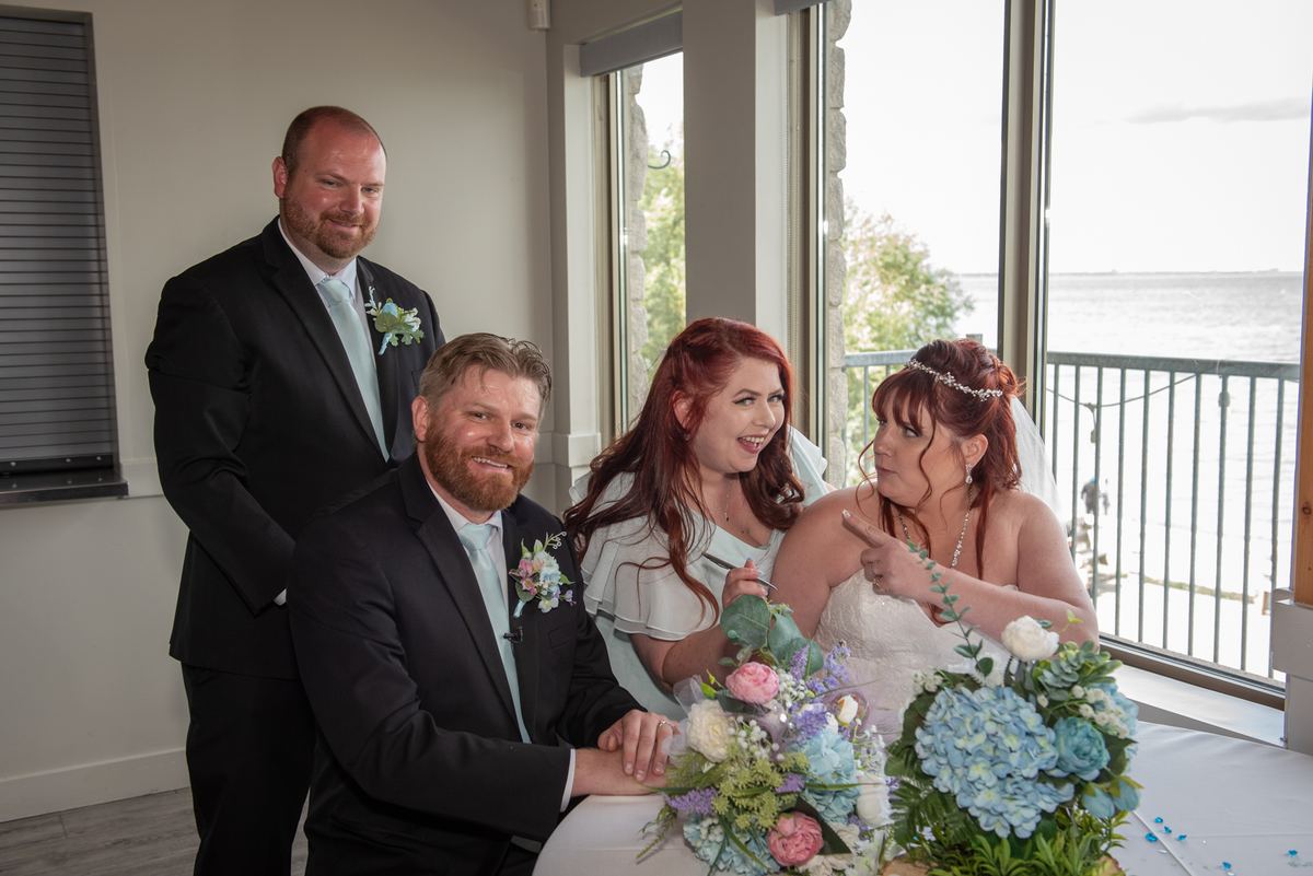 Bride and groom seated at a table near a window overlooking water, with two men in suits behind them.