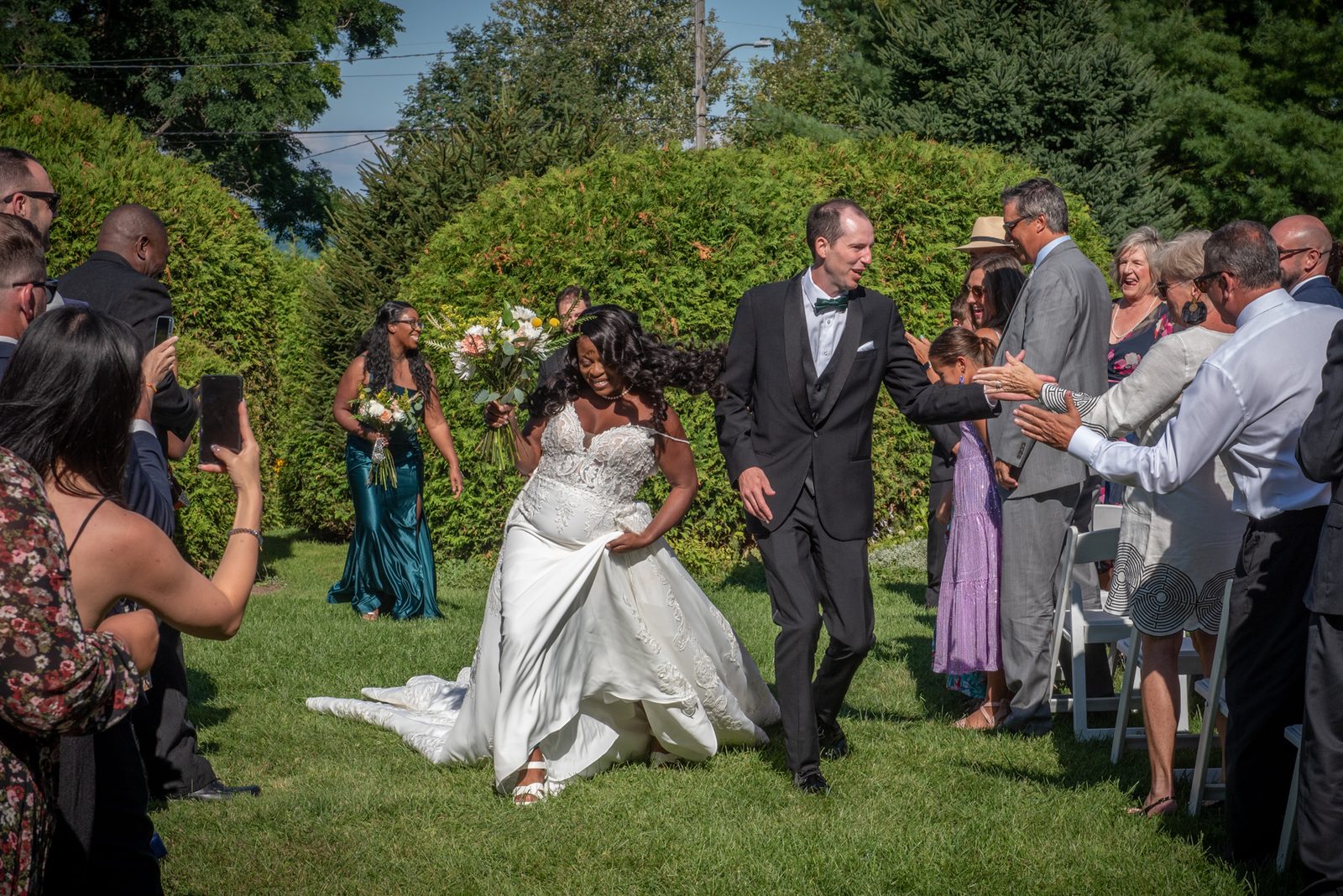 A bride laughs as she walks down a grass aisle with her groom, lifting her train while guests on both sides reach.