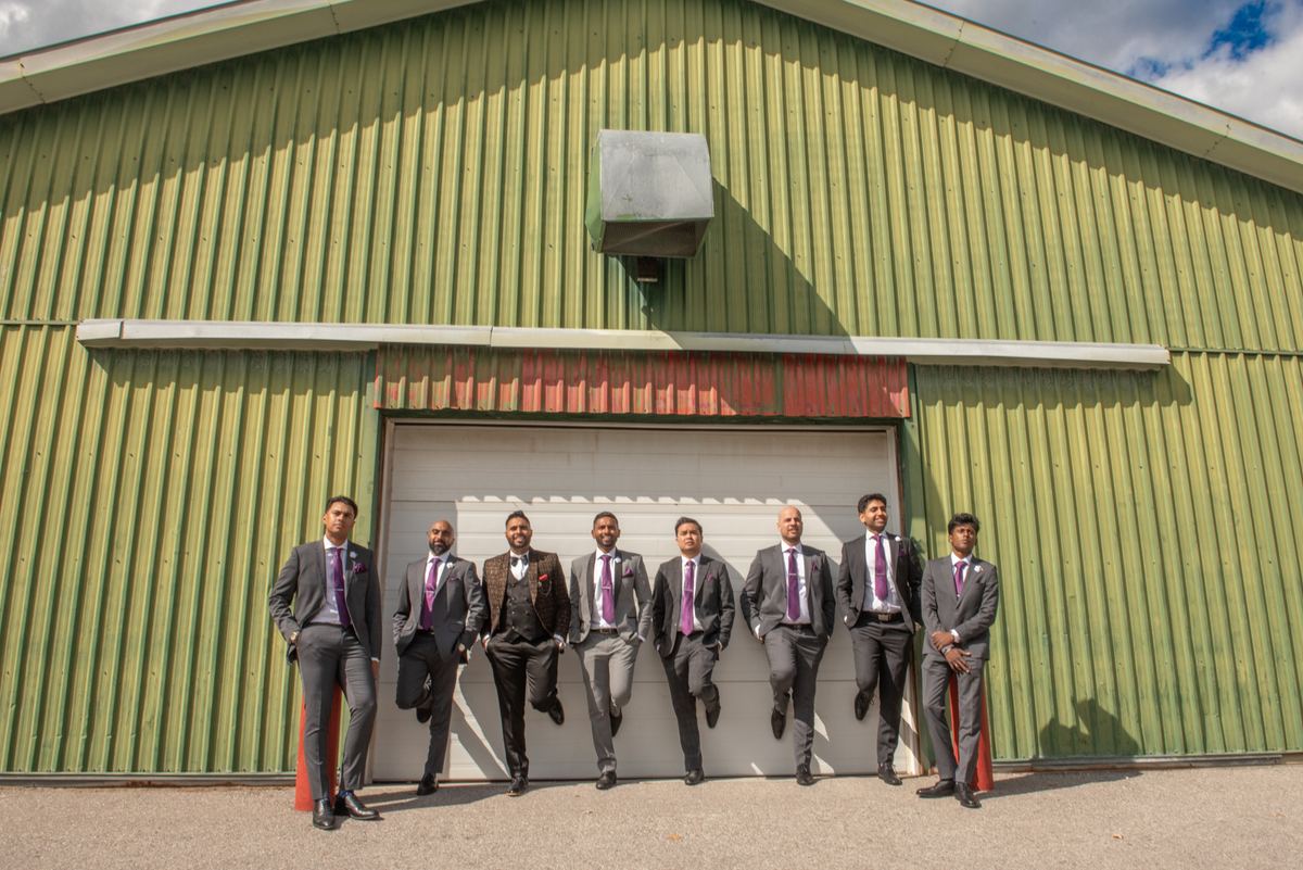 Seven formally dressed men in gray suits and purple ties stand casually before a green metal building.