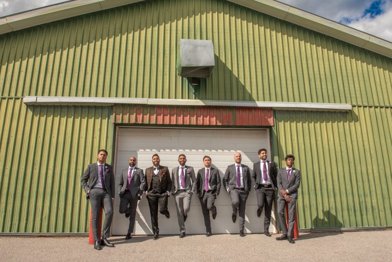 Seven formally dressed men in gray suits and purple ties stand casually before a green metal building.