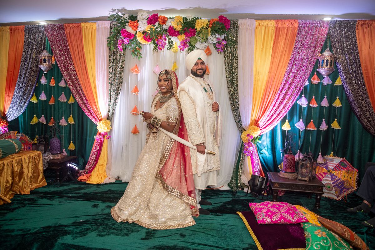 A bride in a sparkling lehenga and a groom in a white sherwani and turban smile before colourful drapes and hanging lanterns.