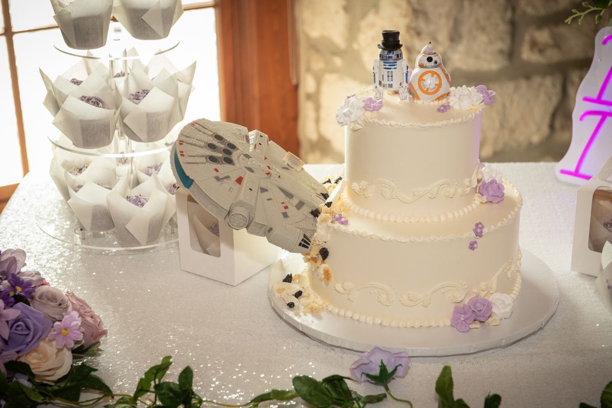 A wedding cake with Star Wars toppers and a Millennium Falcon model, surrounded by cupcakes and purple flowers.