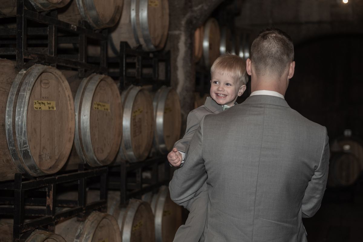 A boy beams as he is held by a man in a suit, standing among wooden barrels in a dimly lit wine cellar.
