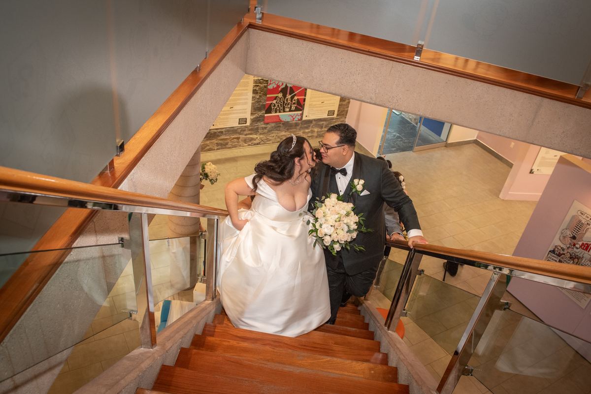Bride and groom kiss while descending a wooden staircase inside a warmly lit building, leaning against the glass railing.