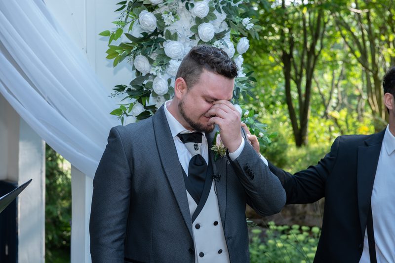 A groom in a gray tuxedo wipes tears under a floral arch as a groomsman comforts him, illuminated by natural light.