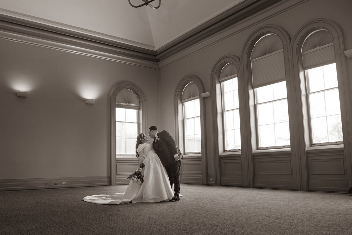 A bride and groom share a tender kiss in an elegant, light-filled room with arched windows and wall sconces.