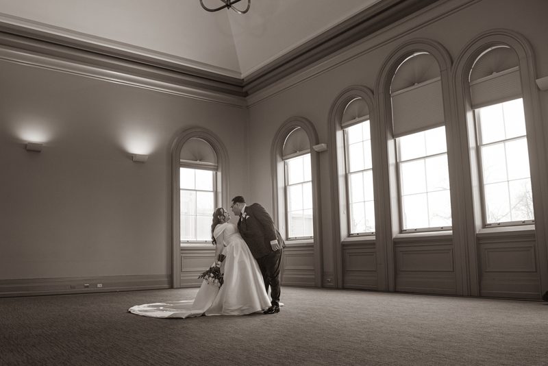 A bride and groom share a tender kiss in an elegant, light-filled room with arched windows and wall sconces.