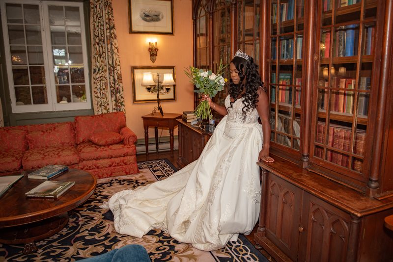 A bride in a white gown and tiara stands by a bookcase in a vintage library with floral decorations.
