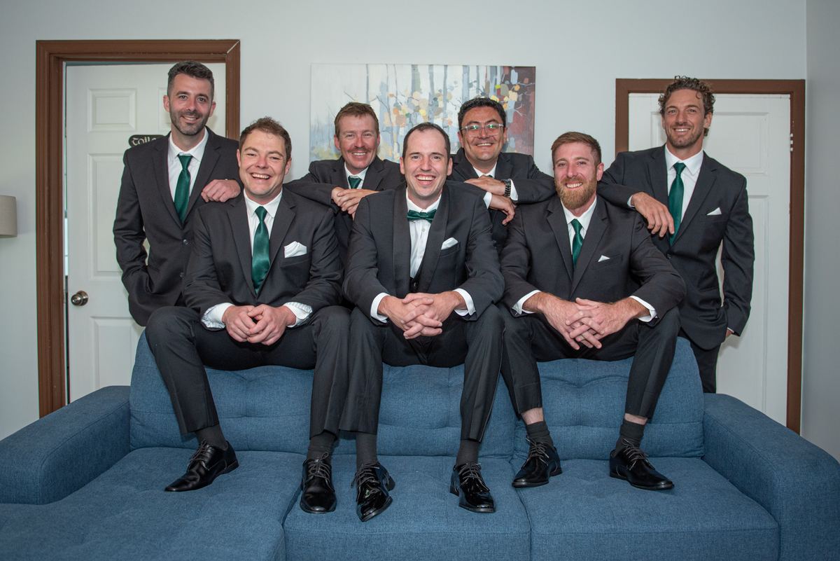 Seven men in dark suits and green ties smile and embrace on a blue couch, posing as the bridal party.