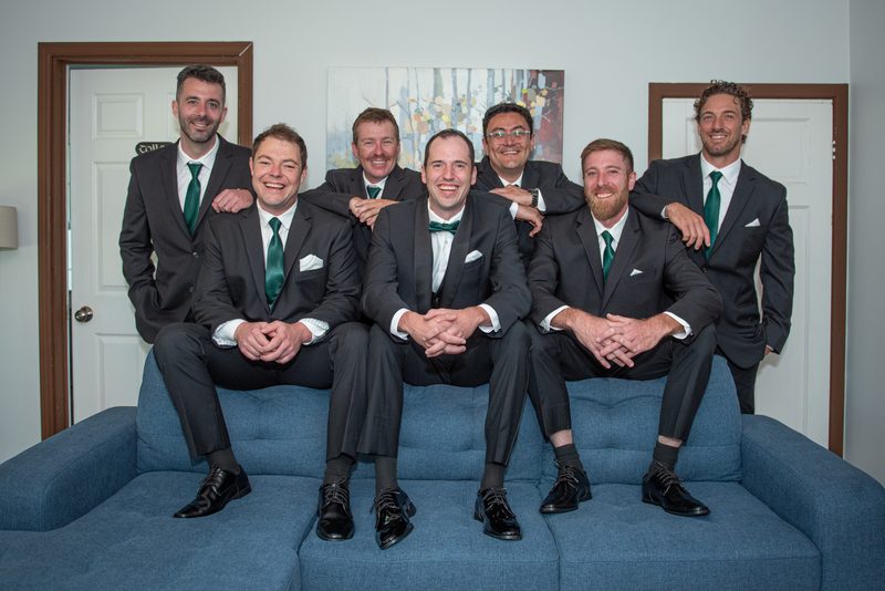 Seven men in dark suits and green ties smile and embrace on a blue couch, posing as the bridal party.