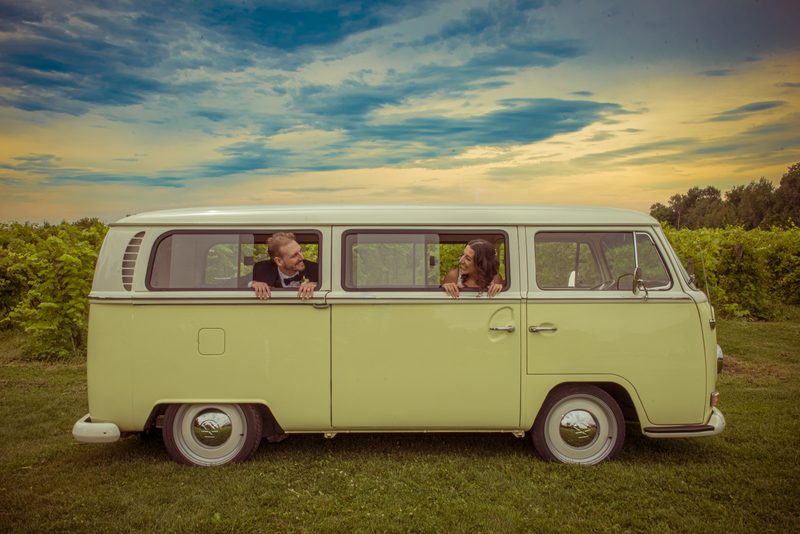 A smiling couple leans out of a vintage green van's windows in a vineyard at sunset, sky glowing with warm hues.