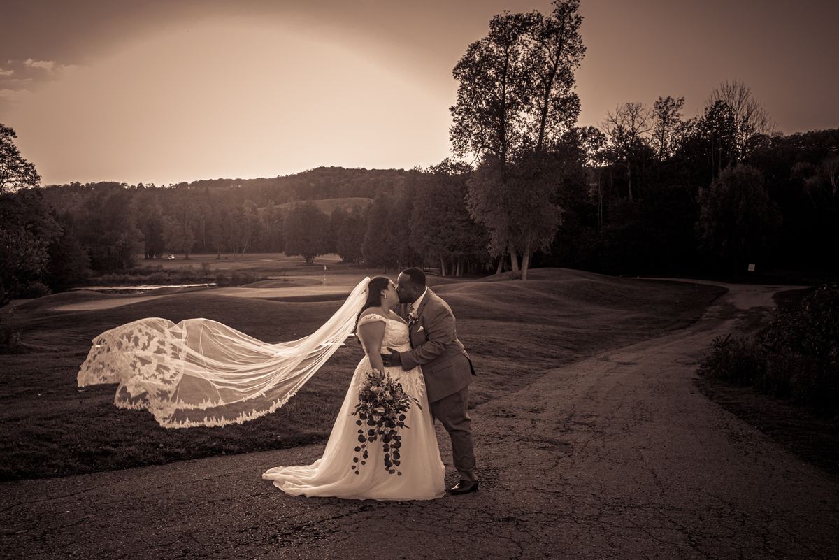 A newlywed couple kisses by a dirt path beside a golf course, the bride's veil billowing in the warm late afternoon breeze.