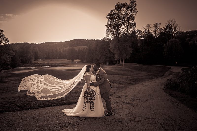 A newlywed couple kisses by a dirt path beside a golf course, the bride's veil billowing in the warm late afternoon breeze.