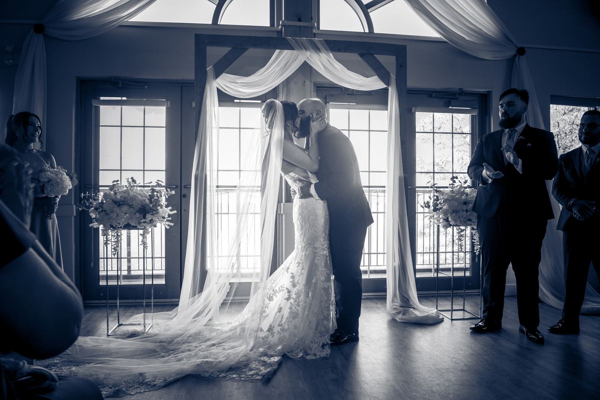A bride and groom kiss under a draped arch at an indoor ceremony, surrounded by guests in formal attire.