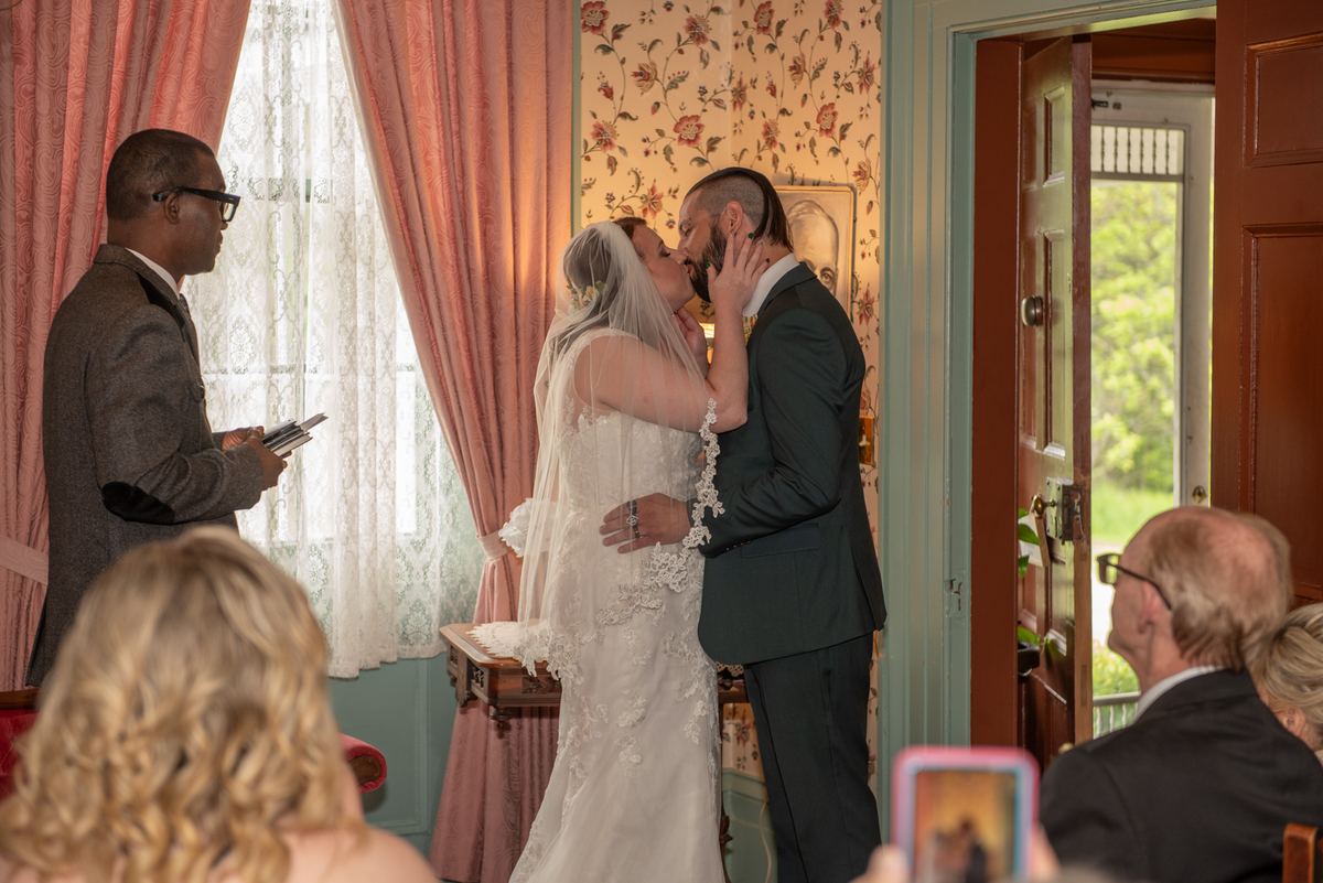 A bride and groom kiss during their ceremony, surrounded by floral wallpaper and pink drapes, with an officiant nearby.