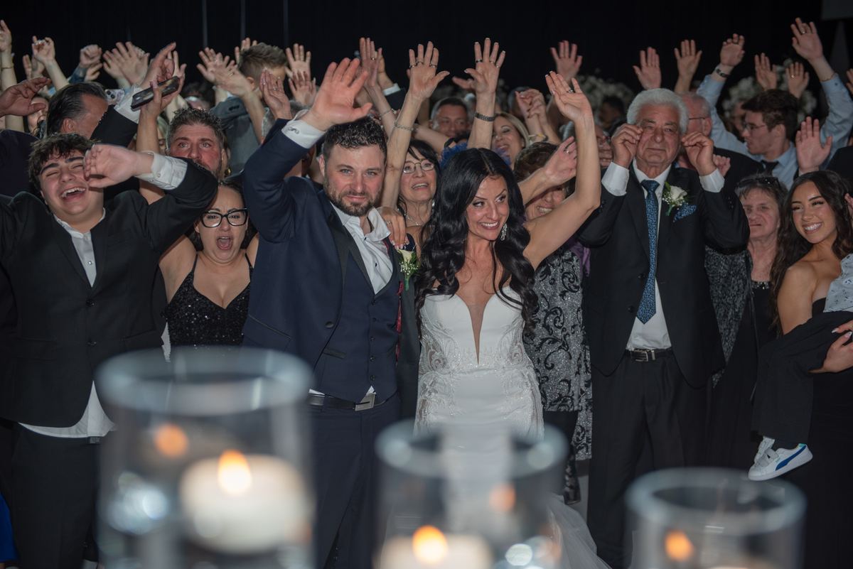 Bride and groom surrounded by cheering guests raising hands in celebration, illuminated by warm candlelight.