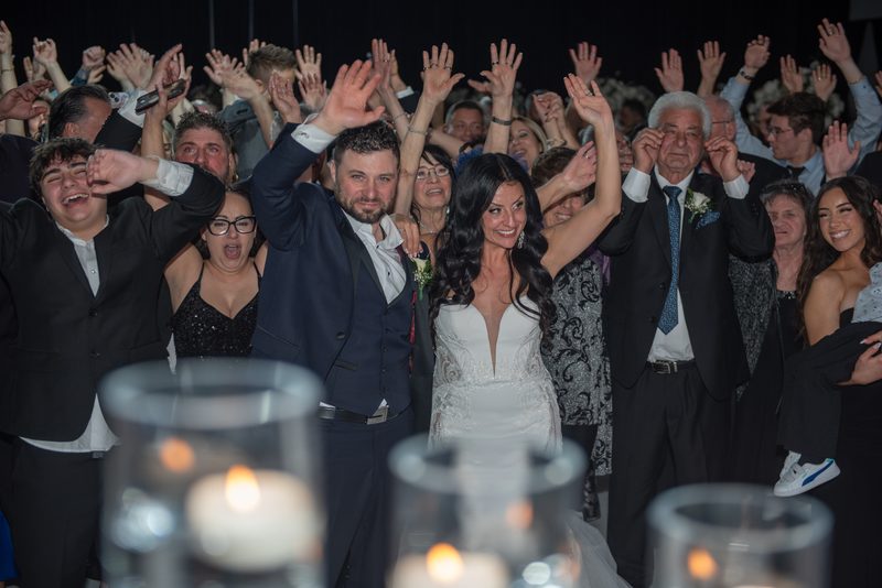 Bride and groom surrounded by cheering guests raising hands in celebration, illuminated by warm candlelight.
