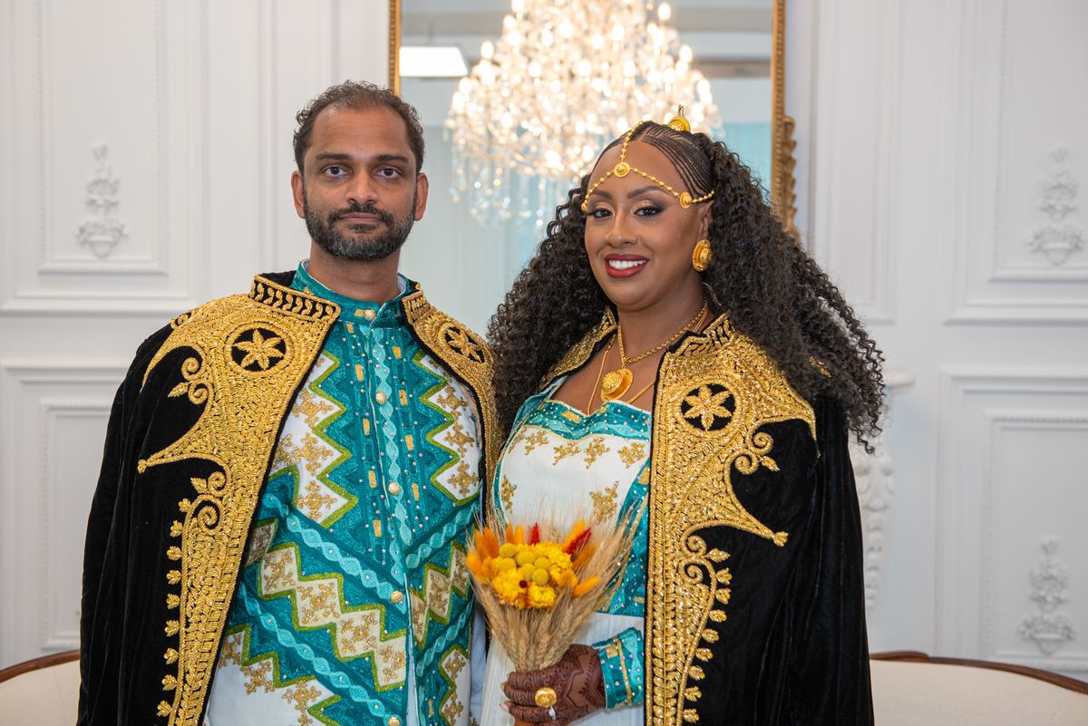 A smiling couple in gold-embroidered jackets and headpieces stands indoors with a bouquet, near a chandelier.