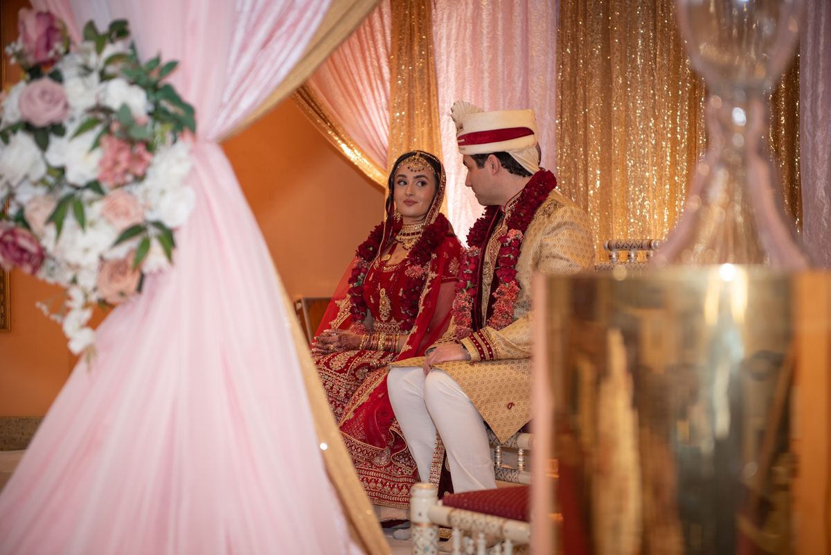 A bride and groom in traditional Indian attire sit together, gazing intimately as golden light filters through curtains.