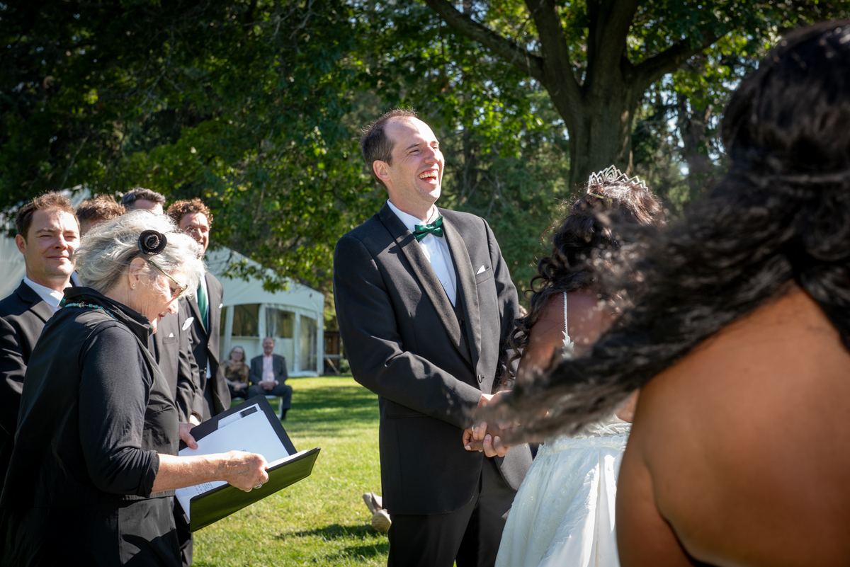 A laughing groom holds his bride's hand during an outdoor ceremony, sunlight filtering through trees.