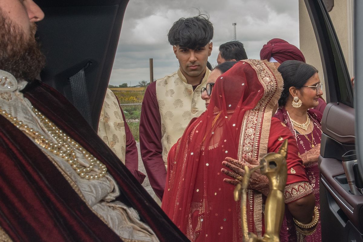 A bride in a red lehenga and dupatta is escorted near a car by family members in traditional attire.