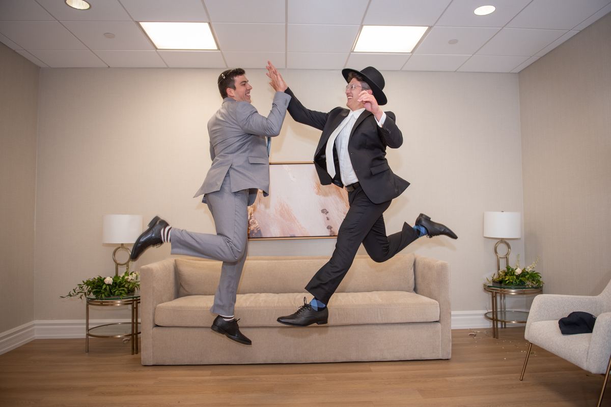 Two formally dressed men jump on a couch, mid-high-five, in a modern lounge with fluorescent lighting.