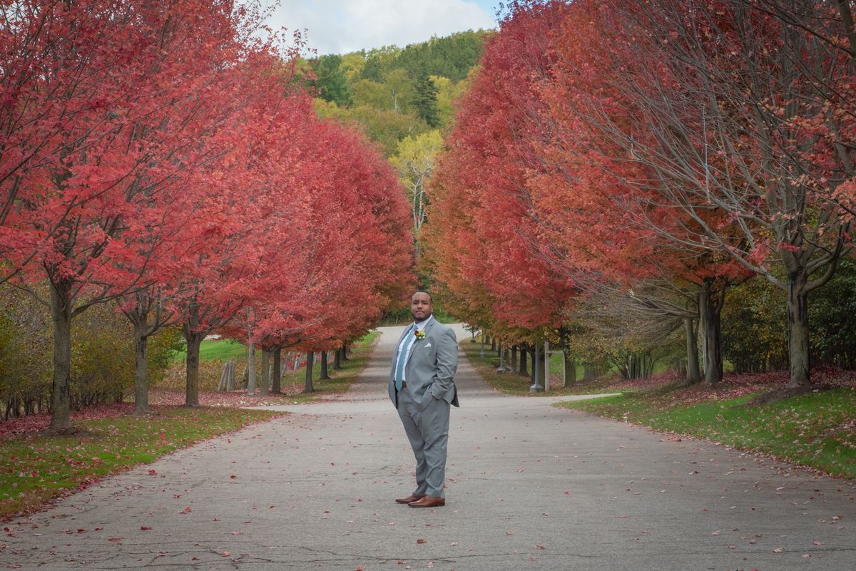 A man in a gray suit stands on a path surrounded by crimson maple trees and fallen leaves, under an overcast sky.